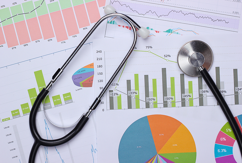 The image shows a stethoscope resting on top of financial documents, including a chart with graphs and numbers.