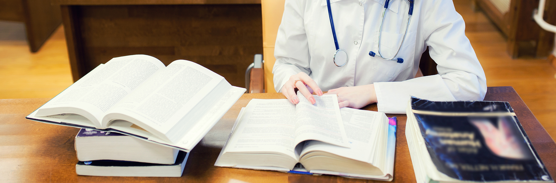 A person seated at a desk with multiple books, wearing a white lab coat, appears to be reading or studying.